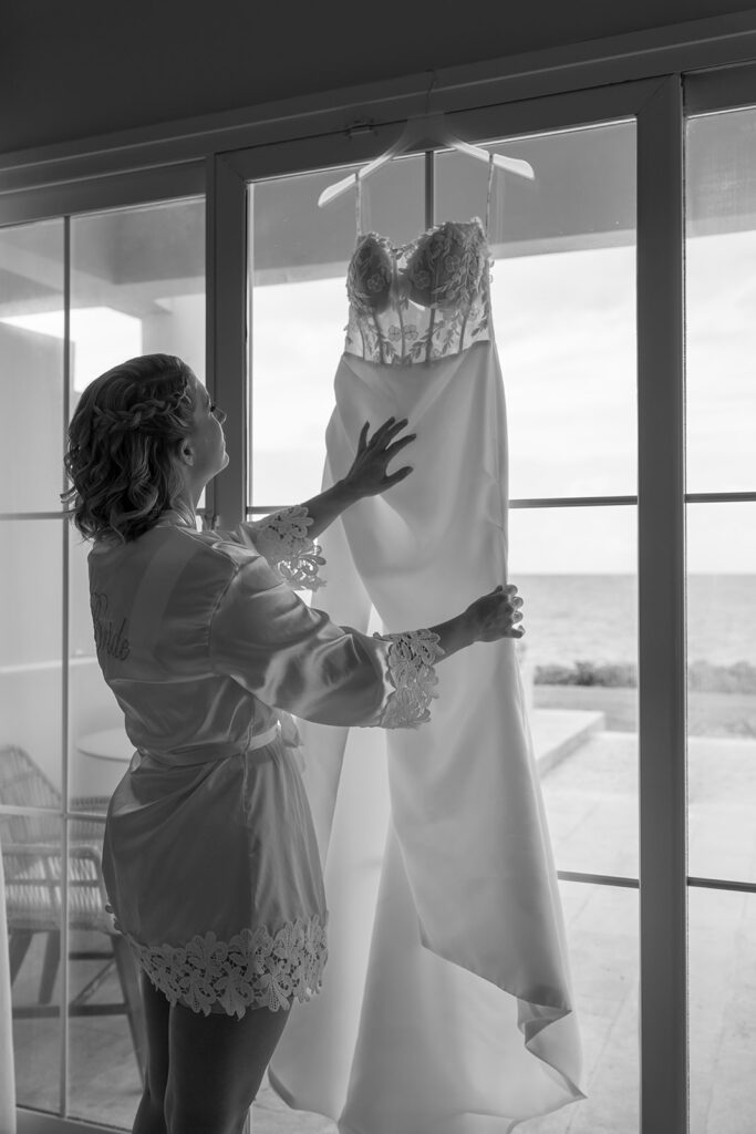 Robe de mariée suspendue devant une fenêtre en bord de mer, préparatifs de mariage à destination