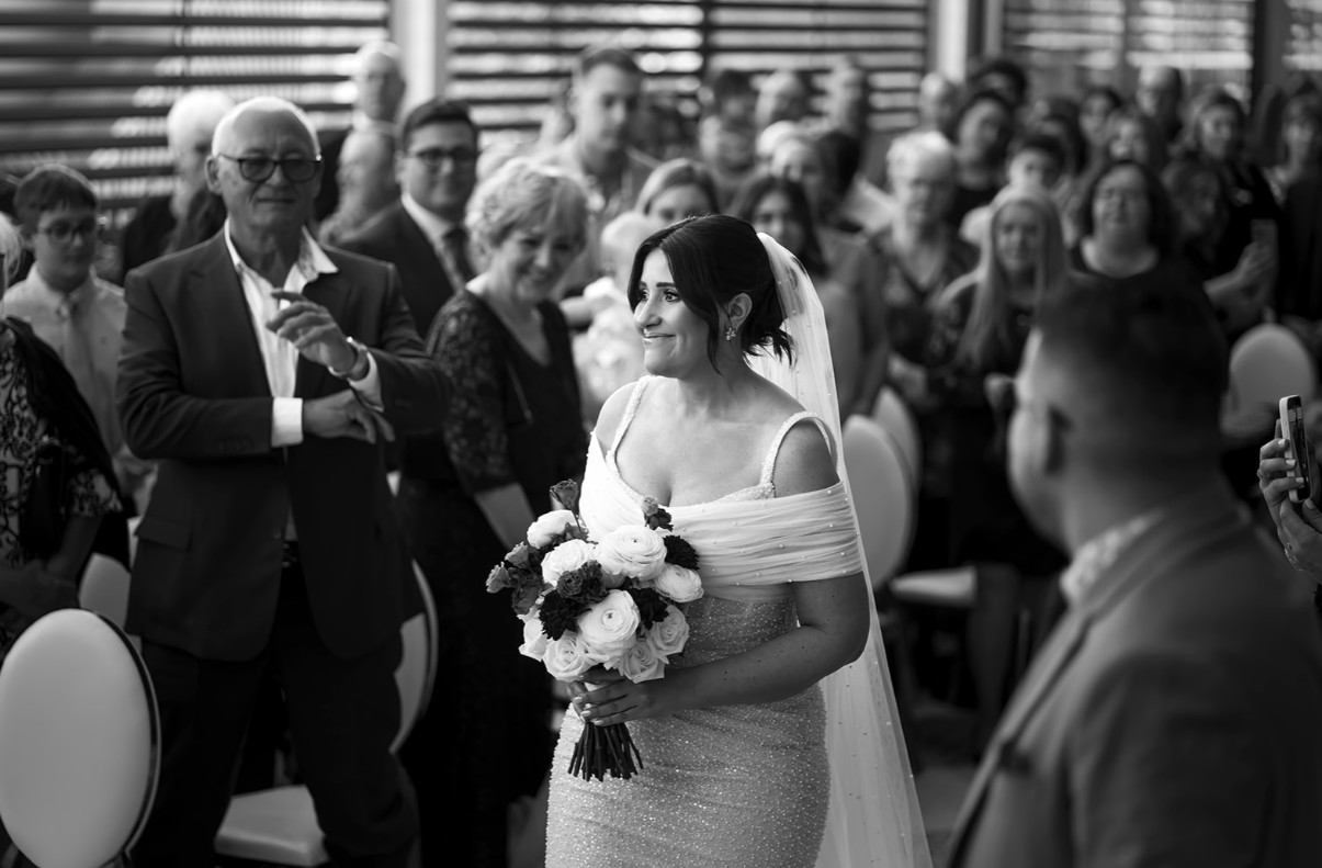 Mariée émue avançant dans l'allée vers son conjoint, accompagnée de son père, entourée de ses invités